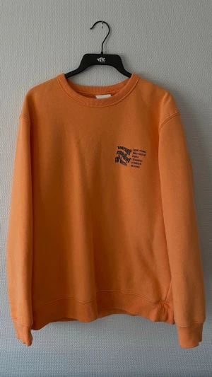 Orange sweatshirt H&M - Orange sweatshirt med svart tryck på bröstet och ryggen. Tröjan har rund hals, ribbade muddar och är gjord i mjukt bomullsmaterial. Perfekt för en avslappnad streetwear-look.