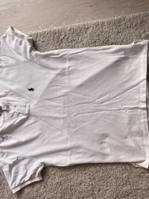 Vit piké från Polo Ralph Lauren M - Säljer en klassisk vit pikétröja från Polo Ralph Lauren i storlek M, custom fit. Tröjan har korta ärmar, krage med två knappar och den ikoniska broderade loggan i marinblått på bröstet. Perfekt för en clean och sportig look.