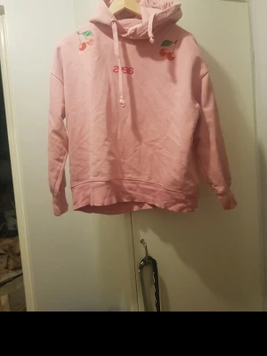 Rosa hoodie med körsbär och text - Säljer en rosa hoodie med huva och dragsko. På bröstet finns små körsbärsbroderier och texten 'sexs' i rött. Hoodien har en avslappnad passform och ribbade muddar vid ärmar och nederkant. Perfekt för en chill och trendig look. Storlek:S/m
