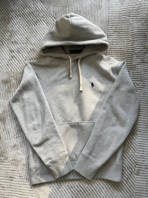 Grå hoodie från Polo Ralph Lauren - Snygg grå hoodie från Polo Ralph Lauren med klassisk huva och dragsko. Tröjan har en broderad marinblå logga på bröstet och en stor magficka framtill. Tillverkad i mjukt bomullsmaterial som känns skönt mot huden. Perfekt för en avslappnad och stilren look. Storlek Xs men passar Xxs, jag på bild är 155cm. Har en fläck som jag tror går bort, sminkfläck. Se bild.