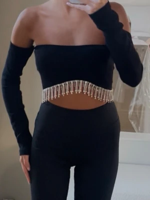 Svart offshoulder topp med pärlor - Trendig svart offshoulder topp med långa ärmar och croppad passform. Toppen har snygga pärldetaljer som hänger längs nederkanten och ger en extra touch. Perfekt för dig som vill sticka ut med en unik och stilren look.