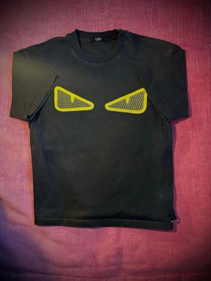 Svart t-shirt med gula ögon - Säljer en svart t-shirt från Fendi med ikoniska gula ögon framtill. T-shirten har rund hals, korta ärmar och är gjord i mjuk bomull. Perfekt för dig som vill sticka ut med en cool och unik design. Skicket är som nytt!
