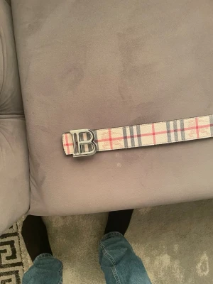 Rutmönstrat bälte med B-spänne - Snyggt bälte från Burberry med klassiskt rutmönster i beige, svart, rött och vitt. Bältet har ett stort metallspänne i form av bokstaven B. Perfekt accessoar för att lyfta din outfit med en ikonisk touch.