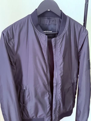 Svart bomberjacka från Peak Performance - Snygg svart bomberjacka från Peak Performance med klassisk ribbad krage, muddar vid ärmslut och nederkant samt dragkedja framtill. Jackan har två sidofickor med dragkedja och är tillverkad i ett lätt, glansigt material som ger en modern look.