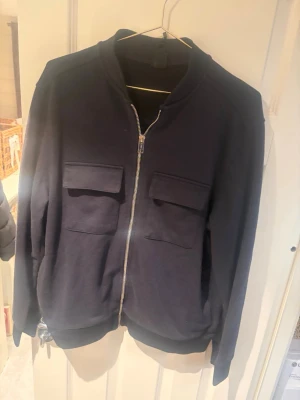 Svart hoodie med dragkedja och fickor - En svart hoodie med dragkedja framtill och två stora fickor med lock på bröstet. Tröjan har ribbade muddar vid ärmslut och nederkant samt en enkel, stilren design utan synligt märke. Tillverkad i mjukt sweatshirtmaterial.