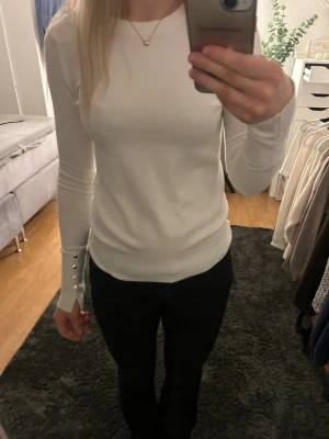 Vit långärmad tröja med gulddetaljer - Säljer en stilren vit långärmad tröja med tight passform och snygga guldfärgade knappar vid ärmsluten. Tröjan har rund halsringning och är gjord i ett mjukt, stretchigt material som sitter skönt på kroppen.