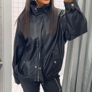 Svart oversized skinnjacka - Snygg svart oversized skinnjacka med hög krage och dragkedja framtill. Jackan har tryckknappar, två fickor med lock och elastisk nederkant för en avslappnad look. Perfekt för dig som gillar streetstyle och vill ha en trendig jacka i garderoben. Den är slutsåld!💕
