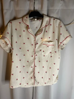 Krämfärgad pyjamas med körsbärsmönster - Söt pyjamas med kortärmad skjorta och shorts i krämfärgat tyg, dekorerad med små körsbär och röda passpoaler. Skjortan har bröstficka och knappar framtill, shortsen har resår i midjan och röd kant. Perfekt för mysiga kvällar.