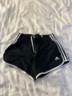 Svarta Adidas shorts med vita ränder - Säljer ett par svarta Adidas shorts med klassiska vita ränder längs sidorna och vit kant. De har elastisk midja med snörning och är perfekta för träning eller chill. Adidas-logga på ena benet och sportig look.