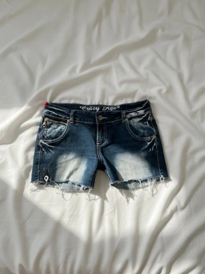 Blå jeansshorts från Crazy Age - Snygga blå jeansshorts från Crazy Age med slitna kanter och broderade detaljer. Shortsen har klassisk femficksdesign, dragkedja och knapp framtill samt coola slitningar och tryck baktill. Perfekta för sommaren och en avslappnad stil.
