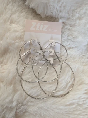 Stora silvriga creoler från Ztiz - Säljer ett set med tre par stora silvriga creoler från Ztiz. Örhängena har en snygg räfflad yta som ger extra glans och attityd. Perfekta för dig som gillar statement-smycken och vill sticka ut lite extra.