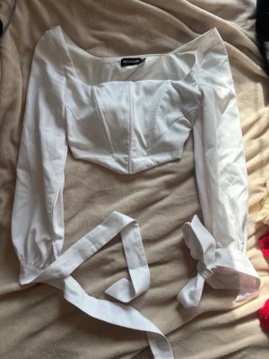 Vit satin cropped topp XS-S – Pretty Little Thing - Vit långärmad cropped topp från Pretty Little Thing i ett satinliknande material 🤍  Toppen har en korsettliknande passform med en liten V-form vid magen som framhäver midjan och höfterna på ett smickrande sätt. Långa snören som knyts vid handlederna ger en extra detalj och dragkedja på sidan.  Storlek EU 34 (XS-S). Jag är 170 cm och brukar ha storlek S/M – toppen sitter tight på mig och går inte riktigt över bysten, så bäst för XS-S.  Använd mycket men väl omhändertagen, i mycket bra skick!✨