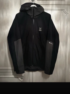 Svart vindjacka från Haglöfs - Snygg svart vindjacka från Haglöfs med grå detaljer på ärmarna och huva. Jackan har dragkedja framtill, två sidofickor med dragkedja och meshfoder på insidan. Tillverkad i slitstarkt material med GORE-TEX-teknologi. Perfekt för blåsiga dagar.