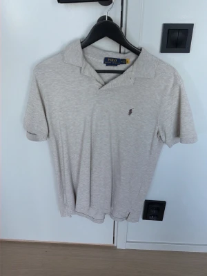 Beige piké från Polo Ralph Lauren - Klassisk beige pikétröja från Polo Ralph Lauren med korta ärmar och liten broderad logga på bröstet. Tillverkad i mjuk bomull och har en stilren krage med knappslå. Perfekt för en clean och avslappnad look.