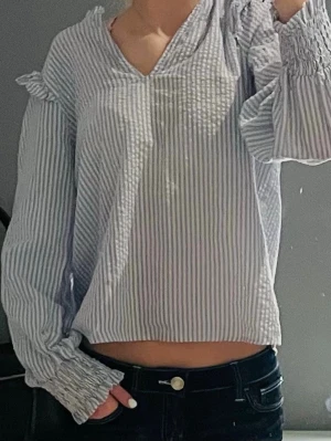 Blårandig blus med volangdetaljer - Snygg blus med blå och vita ränder, volangdetaljer vid axlarna och söt detalj vid i slutet av armen. Blusen har v-ringning och passar till allt möjligt 🤎