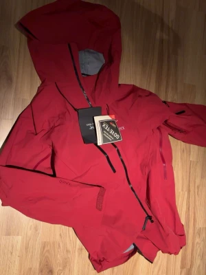 Röd Gore-Tex regnjacka från Arc'teryx - Snygg röd regnjacka från Arc'teryx med justerbar huva och vattentäta dragkedjor. Tillverkad i slitstarkt Gore-Tex-material som håller dig torr även i ösregn. Jackan har flera praktiska fickor och kardborre vid ärmsluten. Perfekt till våren och vandringar, vind och vattentät