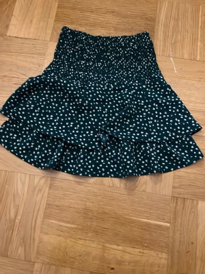 Grön blommig volangkjol - Supersöt kort kjol i mörkgrönt med vitt blommönster och volang (använt 1-2 gånger)OBS M i barn sotorlwk skulle säga att det är ungefär xs i vuxen storlek. 