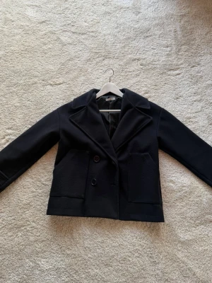 Svart kort kappa från DesignBySi - Stilren marinblå kappa från DesignBySi med dubbelknäppning och stora framfickor. Kortare modell med bred krage och helfodrad insida. Perfekt för dig som gillar minimalistisk och klassisk stil. Materialet ser ut att vara ullblandning. Sällan använd. Storlek xs/s kan uppfattas som större. 