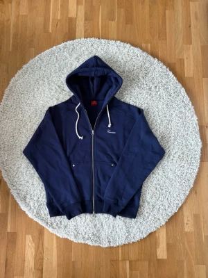 Mörkblå Corteiz hoodie - Mörkblå hoodie från Corteiz  med vita snören och hel dragkedja framtill. Två fickor med knappstängning på framsidan och broderad logga på bröstet. Tillverkad i mjukt bomullsmaterial, perfekt för dig som gillar streetwear! 