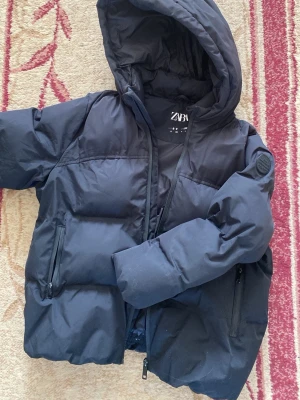 Svart pufferjacka från Zara - Snygg svart pufferjacka från Zara med huva och dragkedja framtill. Jackan har två sidofickor med dragkedja och en patch på ena ärmen. Perfekt för kalla dagar och riktigt skön att bära. Materialet är slitstarkt och håller värmen bra. Min lillebrors jacka som han inte använder längre 💖💖