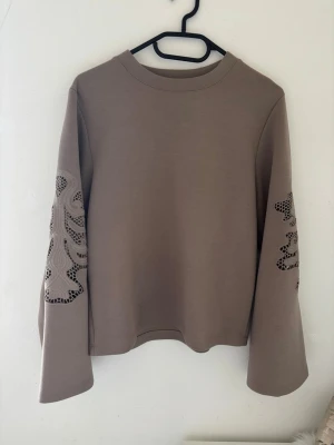 Beige sweatshirt med broderade ärmar från Kappahl  - Säljer en beige sweatshirt med unika broderade detaljer och hålmönster på ärmarna. Tröjan har rund halsringning och vida ärmar som ger en avslappnad look. Perfekt för dig som vill ha något stilrent men ändå speciellt.