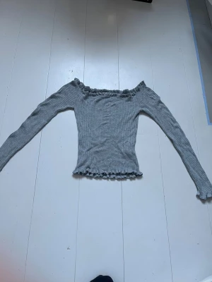 Grå ribbad off shoulder stickad tröja - Trendig grå stickad tröja med ribbad struktur och off shoulder-modell. Tröjan har långa ärmar och volangkant vid halsringning och nederkant för en söt touch. Perfekt för dig som gillar en enkel men snygg stil.