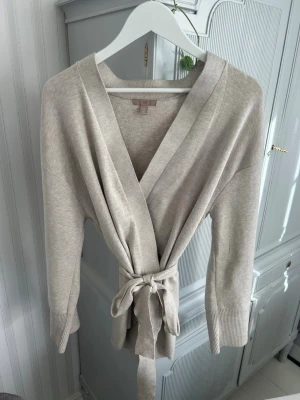 Beige stickad omlottkofta - Beige kofta från H&M i mjukt material. Omlottmodell med knytband i midjan som ger en fin passform. Perfekt både till vardags och lite mer uppklätt.    Använd endast några få gånger och är i mycket fint skick utan fläckar eller skador.