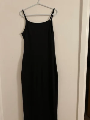 Svart lång klänning med smala axelband - En stilren svart lång klänning med smala axelband och rak passform. Klänningen har minimalistisk design och är tillverkad i ett mjukt, stretchigt tyg som sitter snyggt mot kroppen. Perfekt för dig som gillar enkel och klassisk stil.