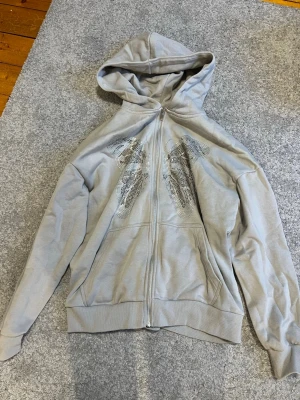 Grå hoodie med strassfjäril - Snygg grå hoodie med dragkedja och huva. På framsidan finns en stor fjäril i silverfärgade strasstenar som ger ett riktigt coolt intryck. Hoodien har fickor framtill och ribbade muddar. Den är väldigt oversized. 