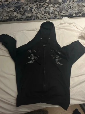 Svart hoodie med bling och ängelmotiv - Svart hoodie med dragkedja och huva, dekorerad med glittrande stenar som bildar texten 'BLING LUCID' och ett coolt ängelmotiv på bröstet. Perfekt för dig som gillar streetstyle och vill sticka ut med lite extra bling. Förlorat några stenar…