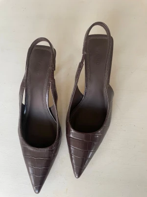 Bruna slingback-pumps från NA-KD - Säljer ett par helt nya (endast provat inomhus få gånger) bruna klackskor från NA-KD med spetsig tå och slingback-rem. Skorna har en klack på ca 5-7cm och ett stilrent krokodilpräglat mönster i konstläder. Perfekta för dig som vill ha en trendig och elegant look. 