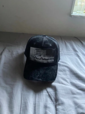 Svart keps med vintage tryck - Säljer en svart truckerkeps med mesh på sidorna och ett stort svartvitt tryck av en sportbil framtill. Kepsen har böjd skärm och justerbar snapback baktill. Perfekt för dig som gillar streetwear och bilar.