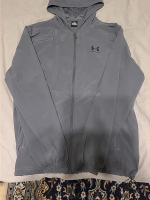 Grå vindjacka från Under Armour - Snygg grå vindjacka från Under Armour med huva och hel dragkedja framtill. Jackan har två sidofickor och elastiska muddar vid ärmsluten. Tillverkad i ett lätt och smidigt material som passar perfekt för blåsiga dagar. Diskret logga på bröstet. Knappast använd!! Priset kan diskuteras.