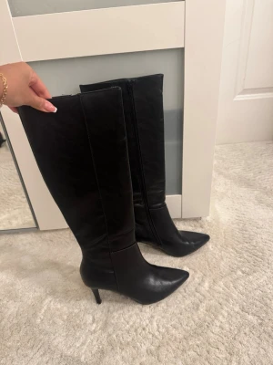Svarta knähöga boots med smal klack från Nelly - Säljer ett par svarta knähöga stövlar med spetsig tå och smal klack. Stövlarna har dragkedja på insidan och är tillverkade i ett skinnliknande material. Perfekta för dig som vill ha en edgy och trendig look.
