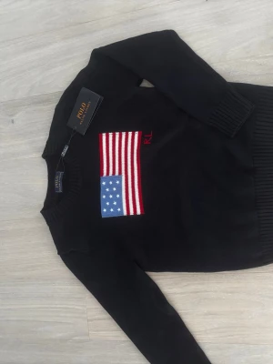 Svart stickad tröja Polo Ralph Lauren - Svart stickad tröja från Polo Ralph Lauren med amerikansk flagga i rött, vitt och blått på bröstet. Tröjan har rund halsringning och ribbade muddar vid ärmslut och nederkant. Märkets logga 'RL' är broderad i rött bredvid flaggan.