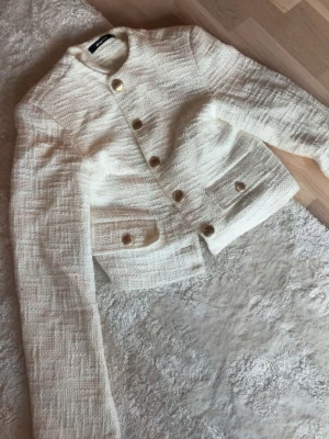 Vit kavajjacka med guldknappar - Stilren vit kavajjacka i strukturerat tyg med dekorativa guldknappar framtill och på fickorna. Jackan har en croppad passform och långa ärmar, perfekt för en trendig och elegant look.