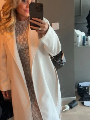 Lång vit kappa med bred krage - Stilren lång vit kappa med bred krage och öppen front. Kappan har en rak passform och är tillverkad i ett mjukt tyg som ger ett elegant intryck. Perfekt för dig som vill ha en klassisk och trendig look.