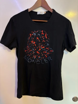 Svart Moncler t-shirt med tryck - Snygg svart t-shirt från Moncler med stort grafiskt tryck i rött och grått på bröstet. Klassisk rund halsringning och korta ärmar. Tillverkad i mjuk bomull som känns skön mot huden. Perfekt för dig som vill ha en stilren men ändå unik look.