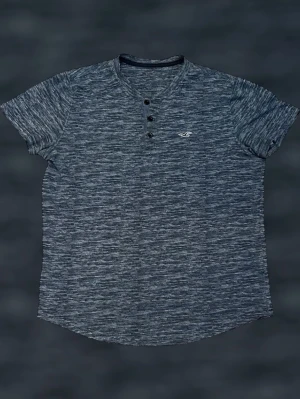  t-shirt från Hollister - Säljer en mörkgrå melerad t-shirt från Hollister med korta ärmar och knappar vid halsen. T-shirten har en liten broderad logga på bröstet och är tillverkad i mjuk bomull. Perfekt för en avslappnad och stilren look.