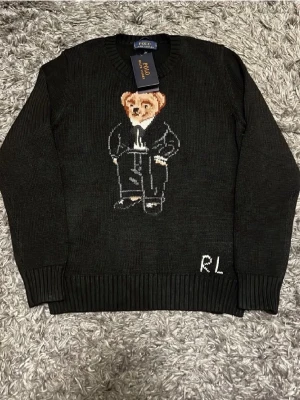 Svart stickad tröja Polo Bear - Svart stickad tröja från Polo Ralph Lauren med ikoniska Polo Bear-motivet framtill. Tröjan har rund halsringning, ribbstickade muddar och RL-broderi vid nederkanten. Perfekt för dig som gillar klassisk och lekfull stil.