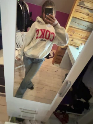 Vit Coca-Cola hoodie med tryck - Säljer en vit hoodie från Coca-Cola med stor röd 'COKE'-logga på bröstet och flera coola tryck på ena ärmen. Hoodien har huva och magficka, samt ribbade muddar. Perfekt för dig som gillar streetwear och vill sticka ut med en ikonisk look.