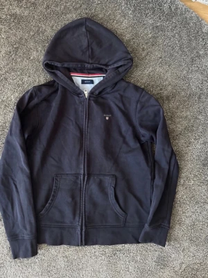 Mörkblå hoodie från Gant - Snygg mörkblå hoodie från Gant med dragkedja framtill och klassisk huva. Hoodien har fickor på magen och en liten Gant-logga broderad på bröstet. Tillverkad i mjuk bomull som känns skön mot huden. Perfekt för en avslappnad och stilren look. Storlek 158/164 13-14 år