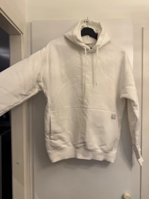 Vit hoodie med huva och magficka - Säljer en stilren vit hoodie med stor huva och dragsnören. Hoodien har en rymlig magficka framtill och ribbade muddar vid ärmslut och nederkant. Perfekt för dig som gillar en enkel och clean look. Materialet känns mjukt och bekvämt. Inga skador eller fläckar