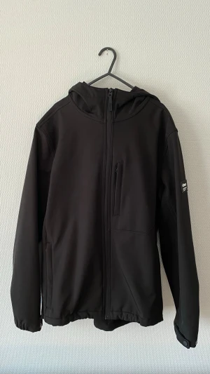 Svart vindjaca Zara - Snygg svart vindjacka från Zara med huva och dragkedja framtill. Jackan har en bröstficka med dragkedja, två sidofickor och elastiska muddar vid ärmsluten. Tillverkad i ett mjukt och smidigt material som står emot vind och vatten.