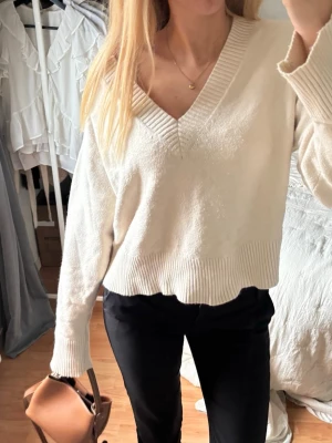Vit stickad V-ringad tröja - Säljer en krämvit stickad tröja med djup V-ringning och bred ribbad kant vid hals, ärmslut och nederkant. Tröjan har en avslappnad passform och långa ärmar, perfekt för lager på lager. Enkel och stilren design utan synligt märke.
