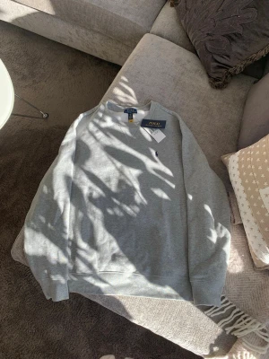 Äkta! Grå sweatshirt från Polo Ralph Lauren NY!  - Snygg grå sweatshirt från Polo Ralph Lauren med klassisk rund halsringning och diskret broderad logga på bröstet. Tröjan har ribbade muddar vid ärmslut och nederkant. Perfekt för en avslappnad och stilren look. Storlek XL barn, 18-20. 
