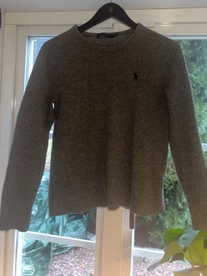 Grå stickad tröja från Ralph Lauren - Mysig grå stickad tröja från Ralph Lauren med rund halsringning och diskret broderad logga på bröstet. Tröjan har långa ärmar och ribbade muddar vid ärmslut och nederkant. Perfekt för lager på lager och enkel att matcha.