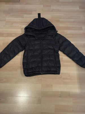 Svart dunjacka med huva - Snygg svart dunjacka med huva och dragkedja framtill. Jackan har quiltad design, två sidofickor med dragkedja och elastiska muddar vid ärmslut och nederkant. Perfekt för kalla vinterdagar och riktigt varm tack vare dunfyllningen.