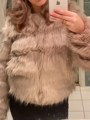 Beige fluffig fuskpälsjacka - Säljer en beige, fluffig fuskpälsjacka med rund halsringning och lång ärm. Jackan har en kort, boxig passform och är tillverkad i mjukt syntetmaterial som ger en lyxig känsla. 