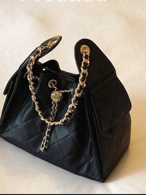 Svart quiltad handväska med guldkedja - Stilren svart handväska i quiltat tyg med en chunky guldfärgad kedja som axelrem och dekorativ gulddetalj framtill. Väskan har en unik form med höga sidor och lock på båda sidorna. Perfekt för dig som gillar trendiga accessoarer med lyxig känsla.